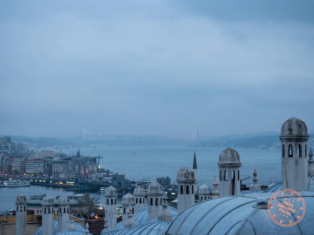 view of rooftops in the foreground and the asia side of istanbul in the background with the bosphorous and bridge at dusk