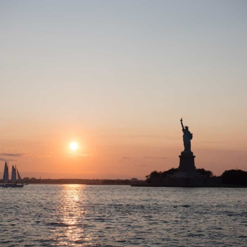 sailboat next to statue of liberty at sunset